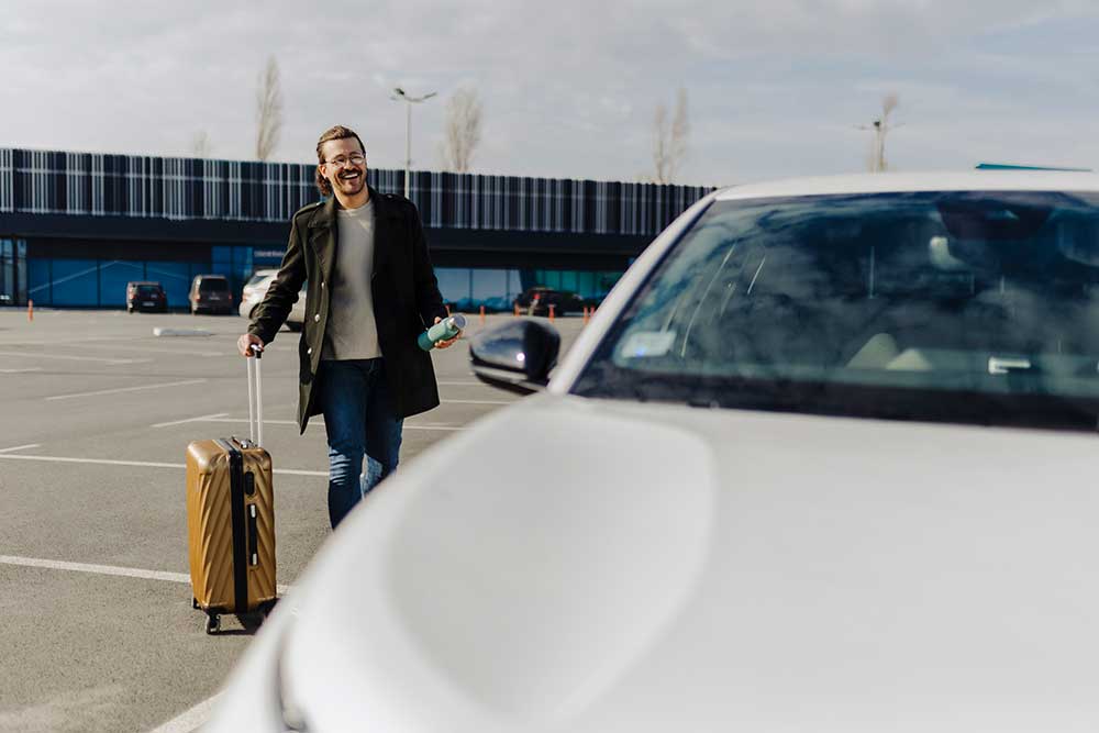 Leende affärsman drar en guldgul resväska på flygplatsens parkeringsplats mot en vit bil