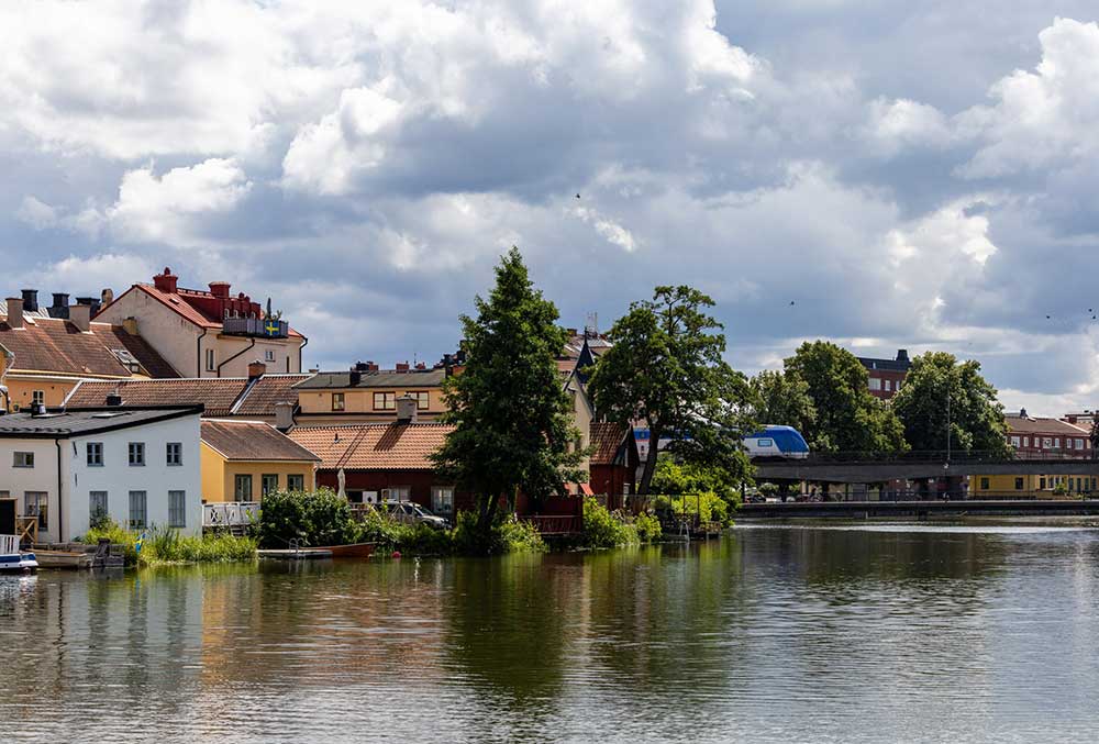 Färgglada bostadshus längs Eskilstunaån med träd och molnig himmel